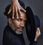 Clarence Brooks posing with a hat in one hand and the other hand spread over the top of his face.